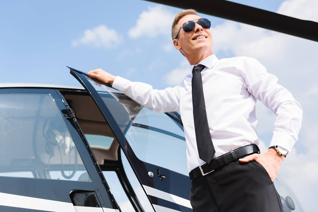 Tiefansicht eines gut aussehenden lächelnden Piloten in formeller Kleidung und Sonnenbrille in der Nähe des Hubschraubers - Foto, Bild