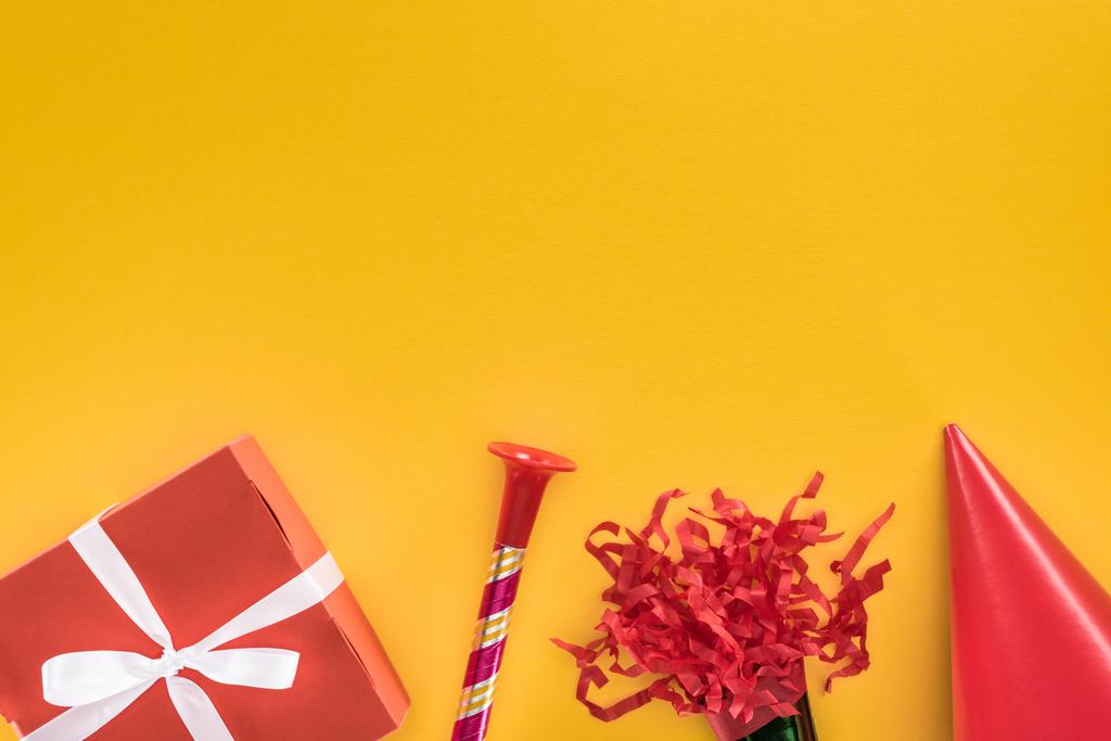 Sarı arka planda kırmızı hediye kutusu, parti kornası ve parti şapkasının üst görünümü - Fotoğraf, Görsel