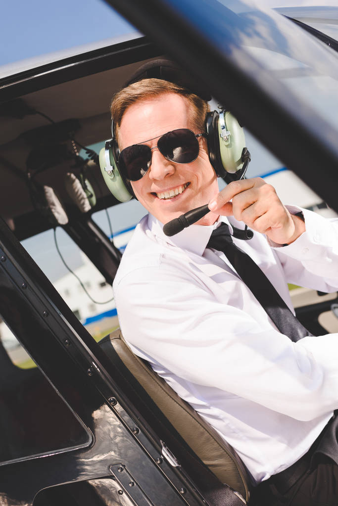 przystojny uśmiechnięty pilot w okulary przeciwsłoneczne i słuchawki siedzi w kabinie śmigłowca - Zdjęcie, obraz