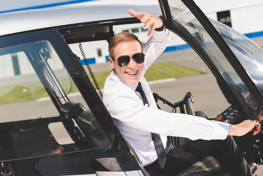 Piloto sonriente con gafas de sol y ropa formal haciendo gestos y sentado en la cabina del helicóptero
 - Foto, imagen