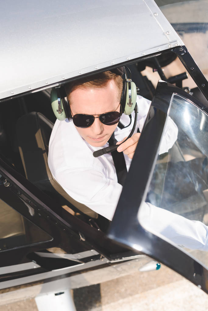 vista superior de Piloto en gafas de sol y auriculares con micrófono sentado en cabina de helicóptero
 - Foto, Imagen