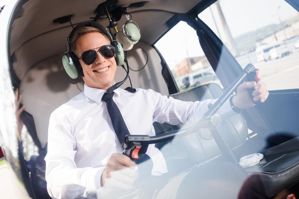 glimlachend piloot in zonnebrillen en headset met microfoon zittend in helikopter cabine en holding Wheel - Foto, afbeelding