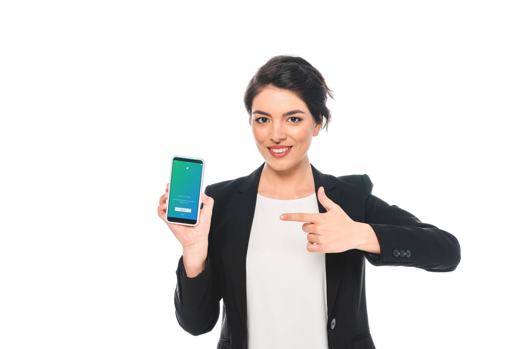 KYIV, UCRANIA - 24 DE ABRIL DE 2019: Mujer de negocios de raza mixta sonriente que señala con el dedo al teléfono inteligente con la aplicación de Twitter en la pantalla aislada en blanco
. - Foto, imagen