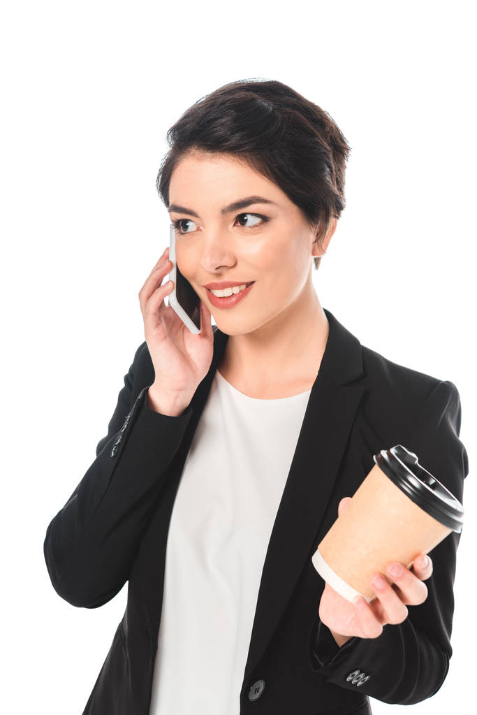 sonriente mujer de negocios de raza mixta hablando en el teléfono inteligente y sosteniendo taza desechable aislado en blanco
 - Foto, Imagen
