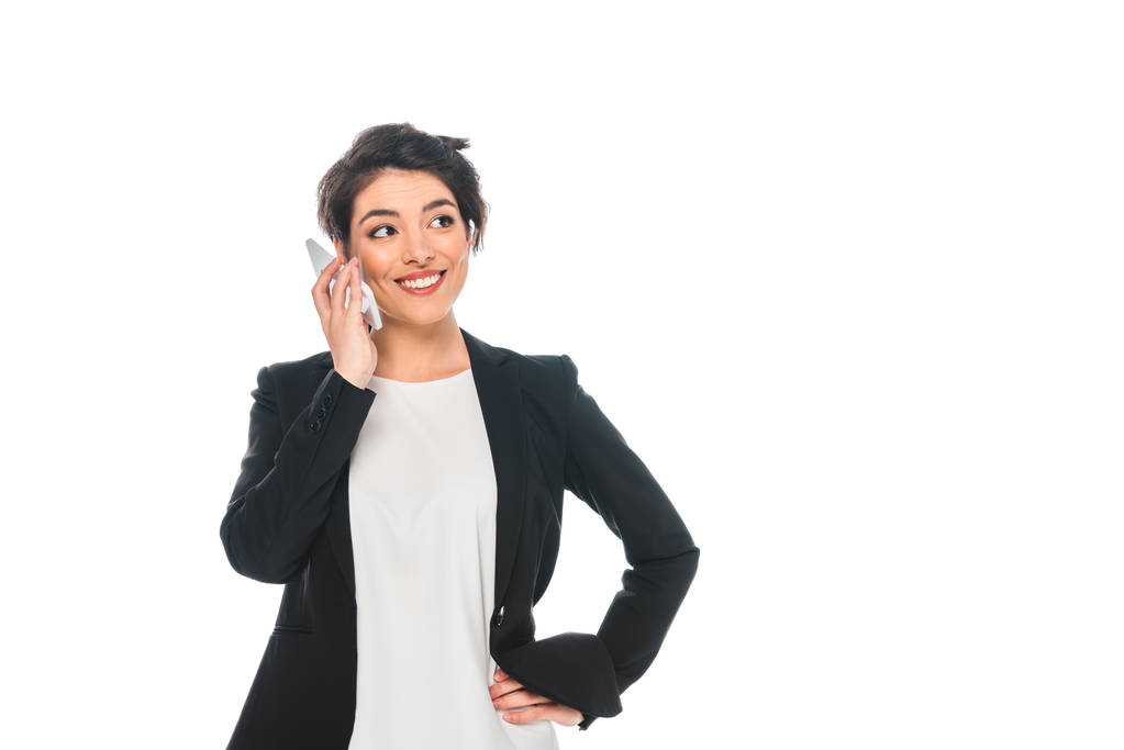 Lächelnde Geschäftsfrau mit gemischter Rasse, die auf dem Smartphone spricht und die Hand auf der Hüfte hält, isoliert auf Weiß - Foto, Bild