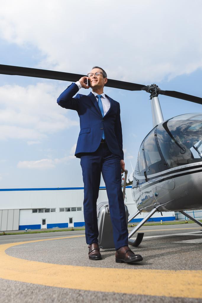 hombre de negocios en ropa formal con maleta hablando en teléfono inteligente cerca de helicóptero
 - Foto, Imagen