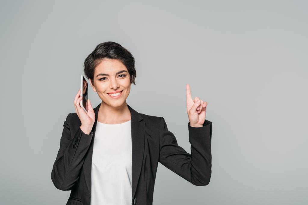 cheerful mixed race businesswoman talking on smartphone and showing idea gesture isolated on grey - Photo, Image