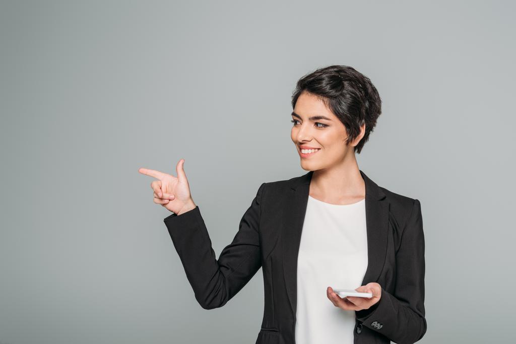 souriant mixte femme d'affaires tenant smartphone et pointant du doigt isolé sur gris
 - Photo, image