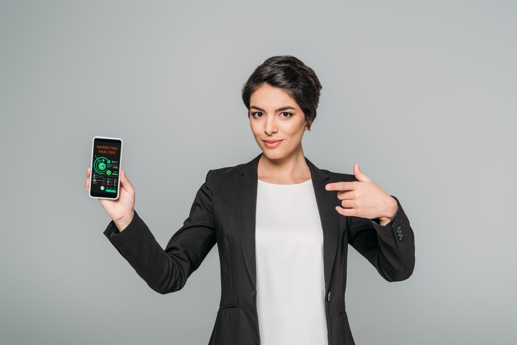 atraktivní smíšená závodní žena ukazující prstem na smartphone s marketingovou analýzou aplikace na obrazovce izolované na šedé - Fotografie, Obrázek