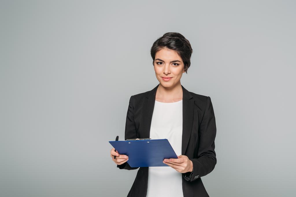 attractive mixed race businesswoman holding clipboard and looking at camera isolated on grey - Photo, Image