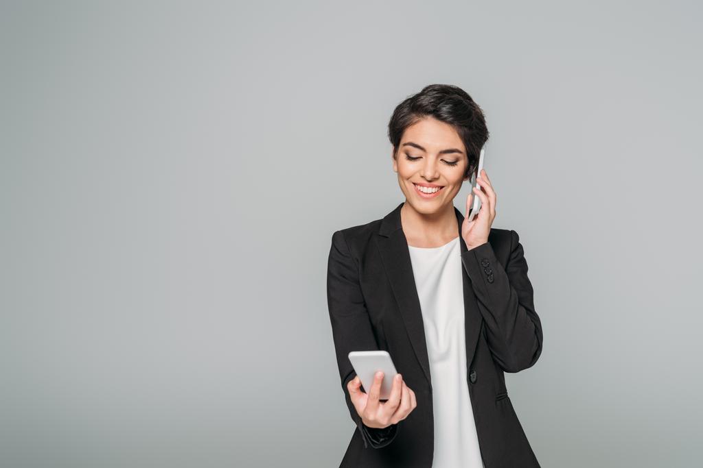 cheerful mixed race businesswoman smiling and using two smartphones isolated on grey - Photo, Image