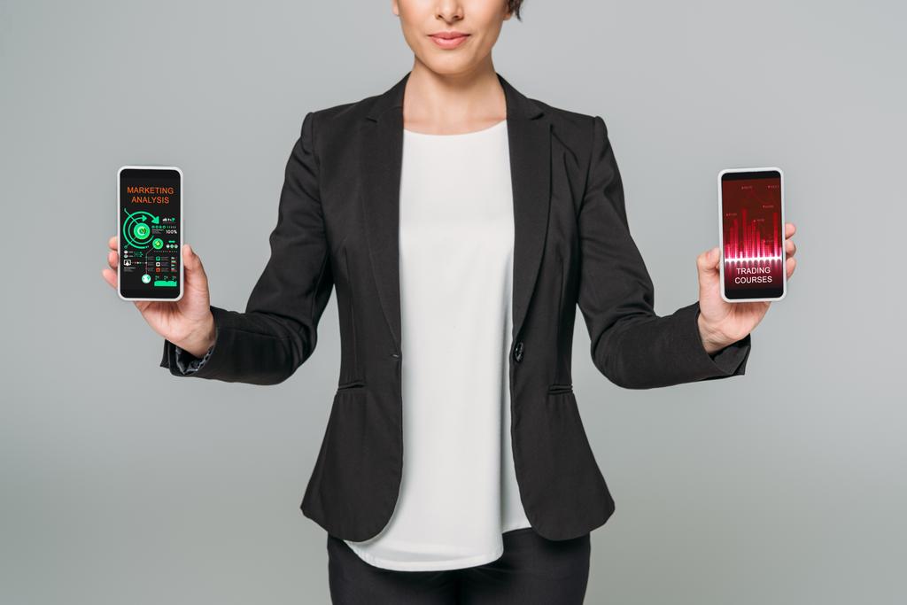 gedeeltelijke weergave van gemengde race zakenvrouw met smartphones met handels cursussen en marketing analyse-apps op het scherm geïsoleerd op grijs - Foto, afbeelding
