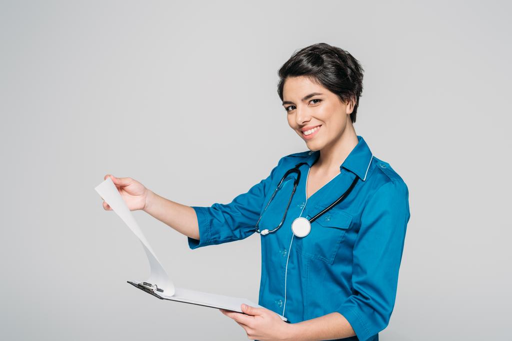 cheerful mixed race doctor holding clipboard with papers and looking at camera isolated on grey - Photo, Image