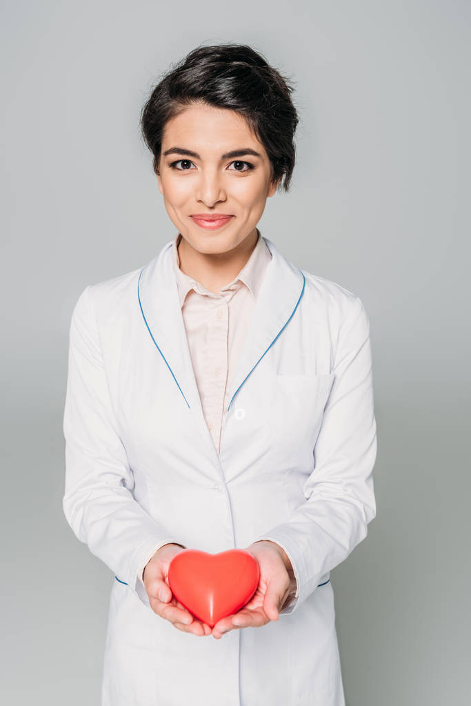 hermosa mezcla de raza médico sosteniendo el modelo de corazón mientras sonríe a la cámara aislada en gris
 - Foto, imagen
