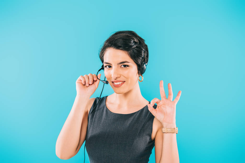 pretty mixed race call center operator in headset showing ok sign while looking at camera isolated on blue - Photo, Image