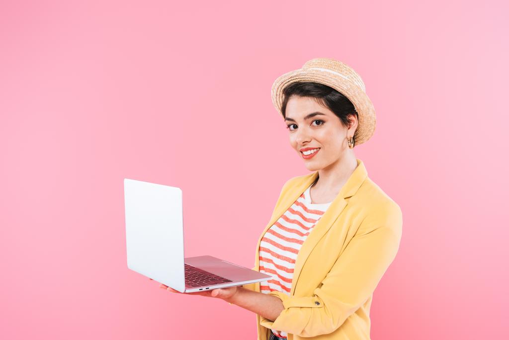 Hübsche Mischlingsfrau mit Strohhut und Laptop, während sie isoliert auf rosa in die Kamera lächelt - Foto, Bild