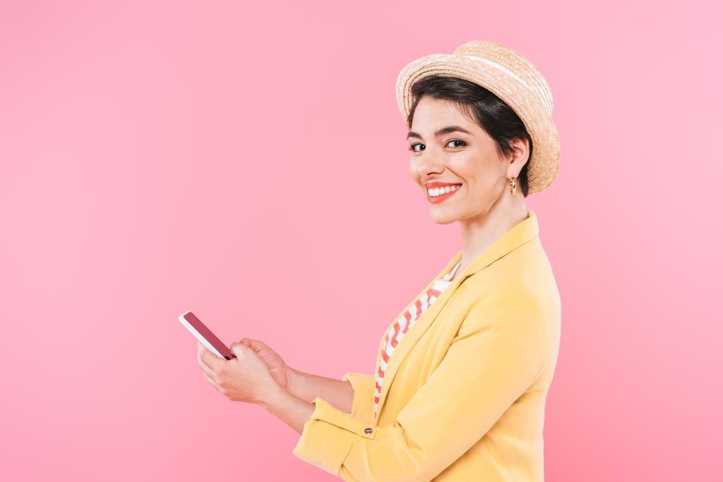 vrolijke mixed race vrouw met behulp van smartphone en glimlachend op camera geïsoleerd op roze - Foto, afbeelding