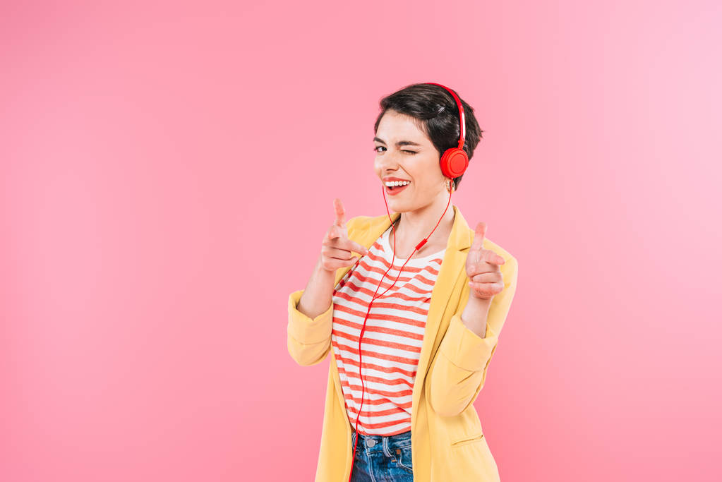 Veselá smíšená žena naslouchající hudbě ve sluchátkách, ukazování prsty a mrknutí na kameru izolované na růžovém - Fotografie, Obrázek