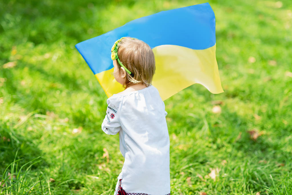 Kind draagt fladderende blauwe en gele vlag van Oekraïne in het veld. Oekraïense Onafhankelijkheidsdag. Vlaggendag. Grondwettelijk dag. Meisje in traditioneel borduurwerk met vlag van Oekraïne. - Foto, afbeelding