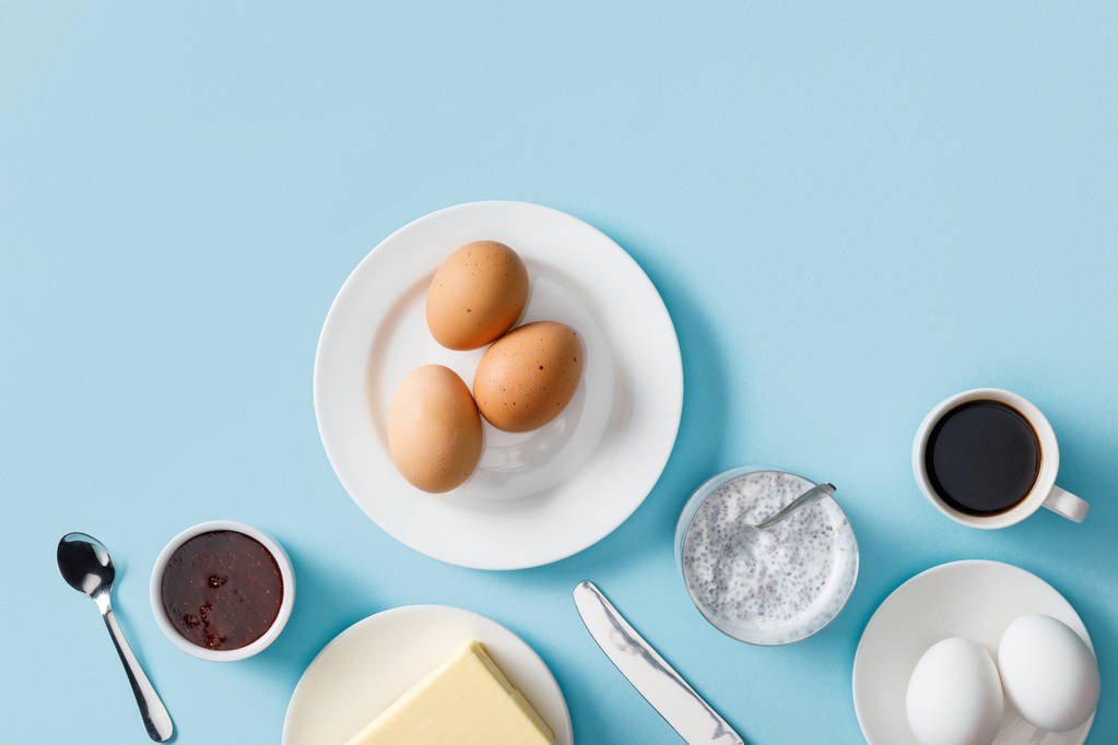 Draufsicht auf frisch gekochte Eier, Butter, Marmelade auf weißen Tellern, Joghurt mit Chiasamen, Kaffee, Löffel und Messer auf blauem Hintergrund - Foto, Bild