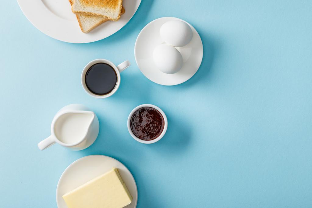 vista superior de huevos frescos, mermelada, mantequilla, café, leche y dos tostadas sobre platos blancos sobre fondo azul
 - Foto, Imagen