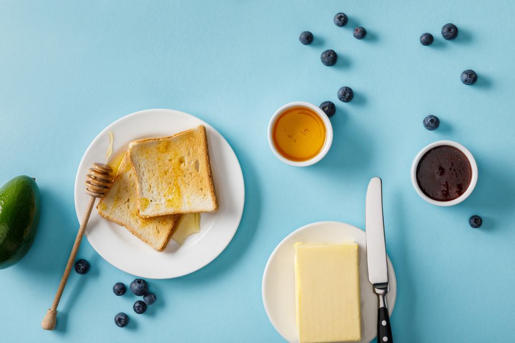 アボカド、バター、蜂蜜、ジャム、散らばったブルーベリー、青い背景の白いプレートに2つのトーストのトップビュー - 写真・画像