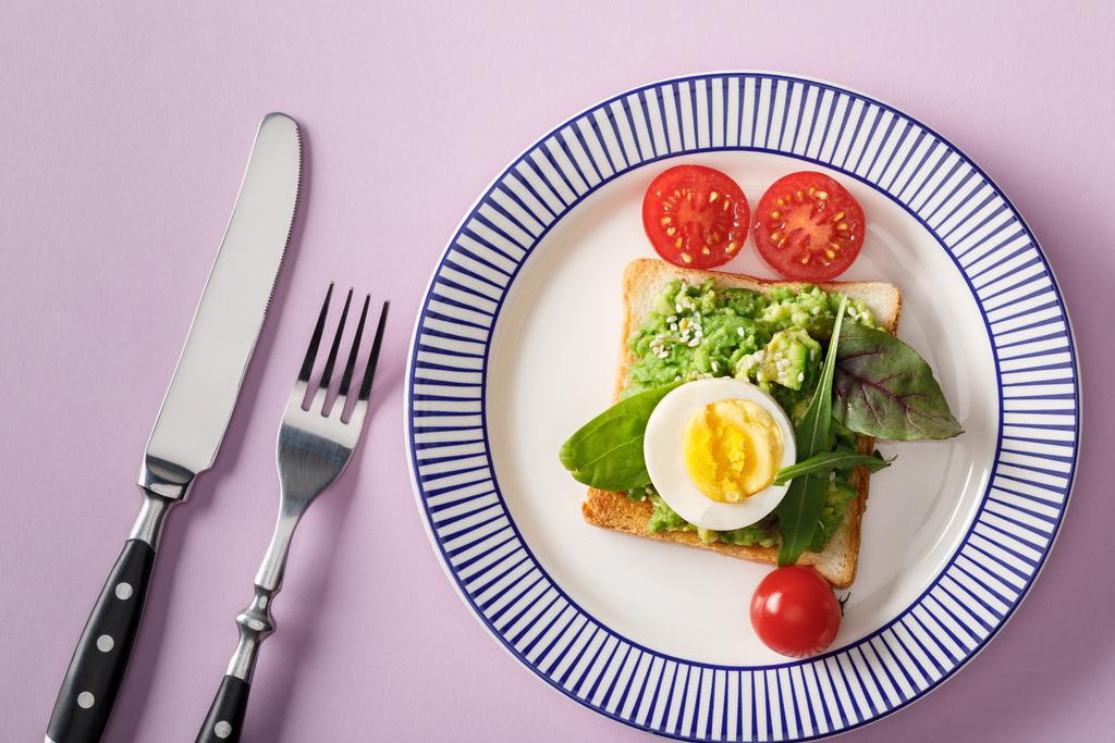 top view of toast with guacamole, boiled egg, spinach, cherry tomatoes, fork and knife on ornamental plate on violet background  - Photo, Image