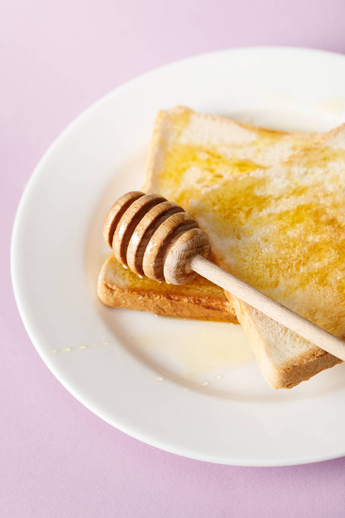 close up view of toasts with honey and wooden dipper on white plate on violet background - Photo, Image