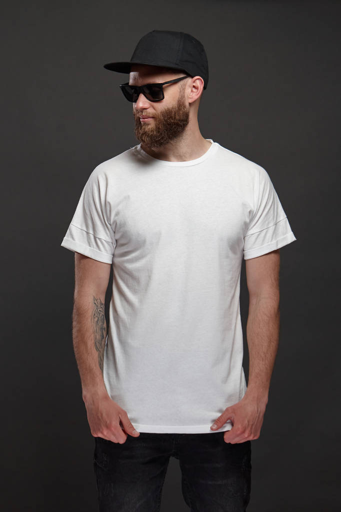 白い空のTシャツとあなたのロゴやデザインのためのスペースを持つ野球帽を身に着けているひげを持つヒップスターハンサムな男性モデルカジュアルな都市スタイルで - 写真・画像