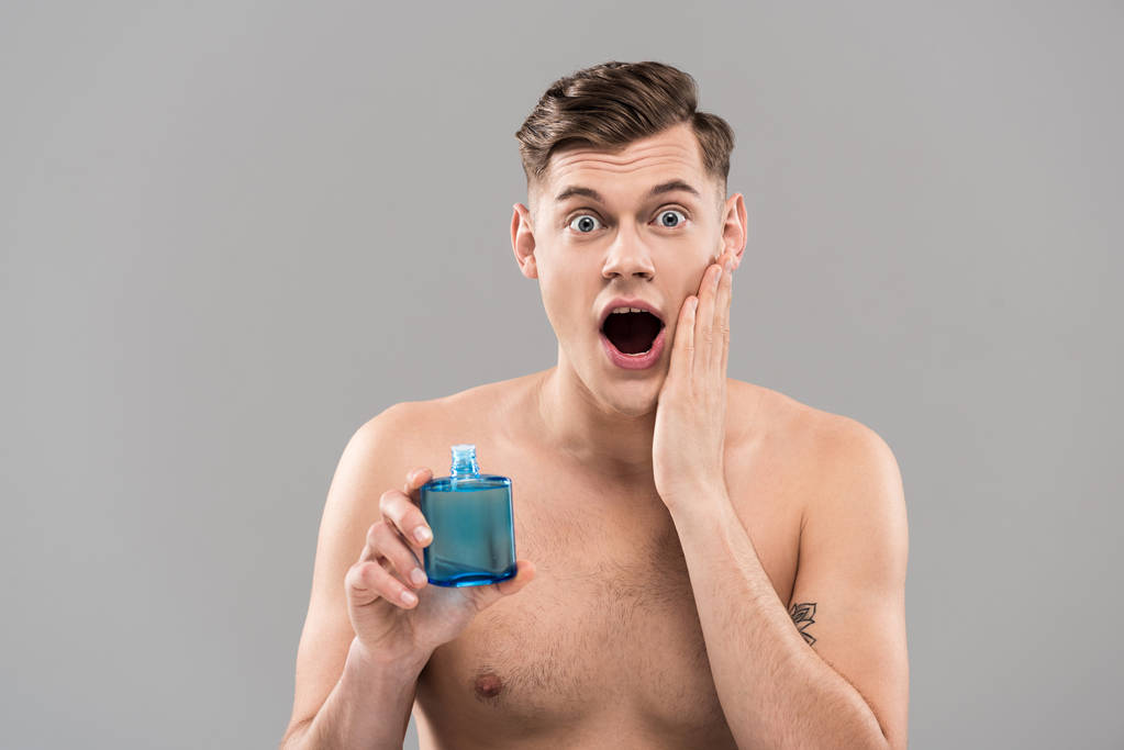 μπροστά όψη του σοκαρισμένος γυμνός νεαρός άνδρας κρατώντας μπουκάλι και εφαρμόζοντας λοσιόν στο πρόσωπο απομονώνονται σε γκρι - Φωτογραφία, εικόνα
