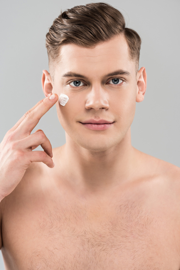 Vorderseite des nackten jungen Mannes, der Gesichtscreme isoliert auf grau aufträgt - Foto, Bild