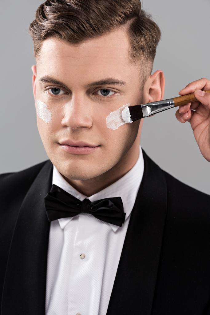 visão recortada de cosmetologista aplicando creme no rosto e homem em desgaste formal isolado em cinza
 - Foto, Imagem