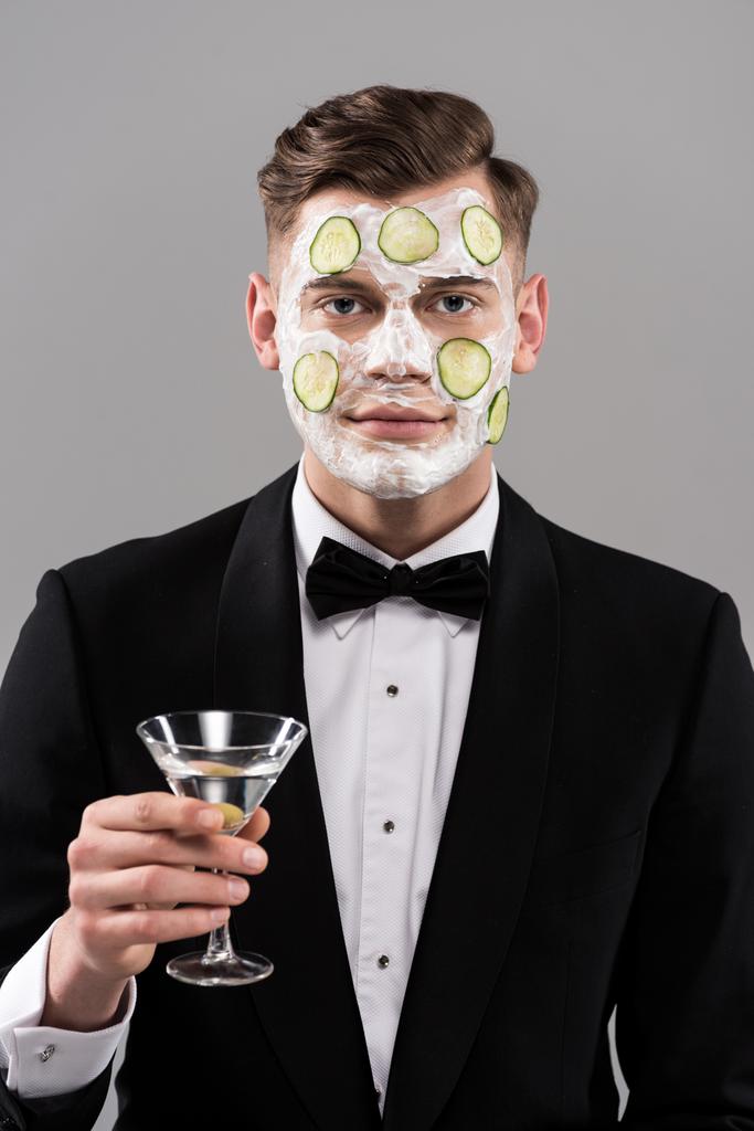 joven en ropa formal con máscara facial de pepino sosteniendo vaso de cóctel aislado en gris
 - Foto, Imagen