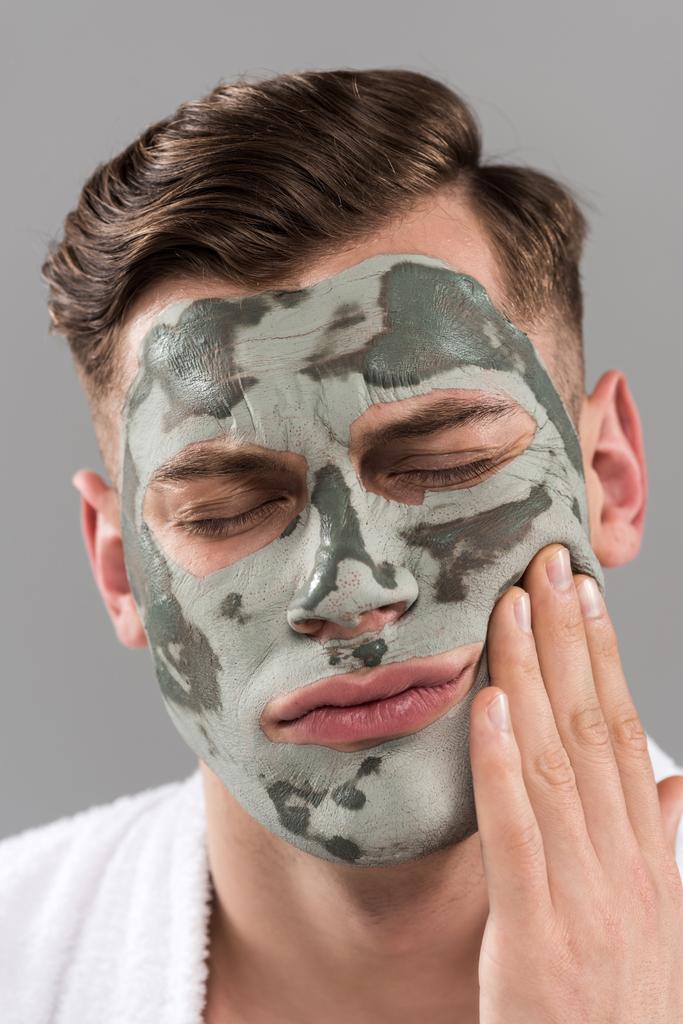 displease jonge man met klei masker aanraken gezicht geïsoleerd op grijs - Foto, afbeelding
