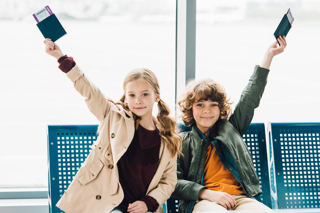 счастливые дети, сидящие на синих сиденьях, держащие паспорта и билеты в зале ожидания в аэропорту
 - Фото, изображение