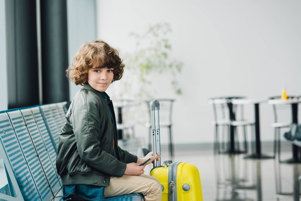 Chłopiec siedzi na niebieskim siedzeniu, trzymając paszport i żółty walizka i patrząc na kamerę w poczekalni na lotnisku  - Zdjęcie, obraz