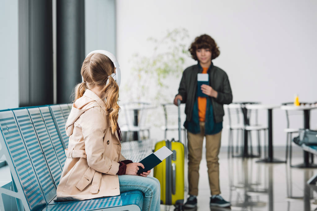 селективное внимание ребенка-подростка, смотрящего на мальчика, стоящего с паспортом и желтым чемоданом в зале ожидания
 - Фото, изображение