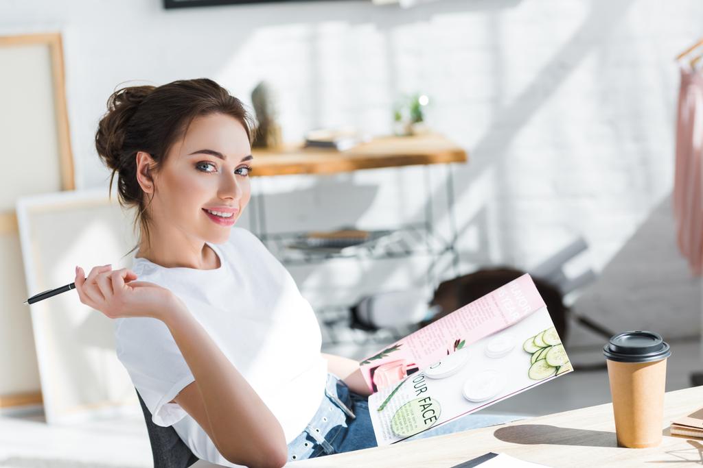 весела жінка в білій футболці тримає журнал і ручку біля паперової чашки на столі
  - Фото, зображення