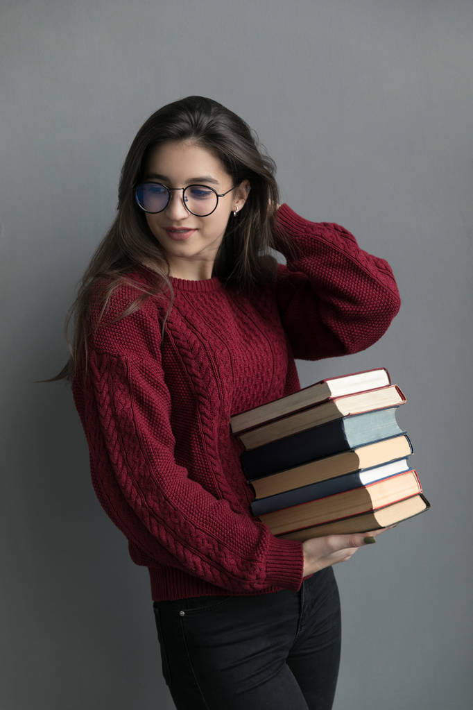 Крупним планом дівчина з тече волоссям, на сірому фоні тримає книги в руках, позує. Носіння бордовий светр і окуляри
. - Фото, зображення