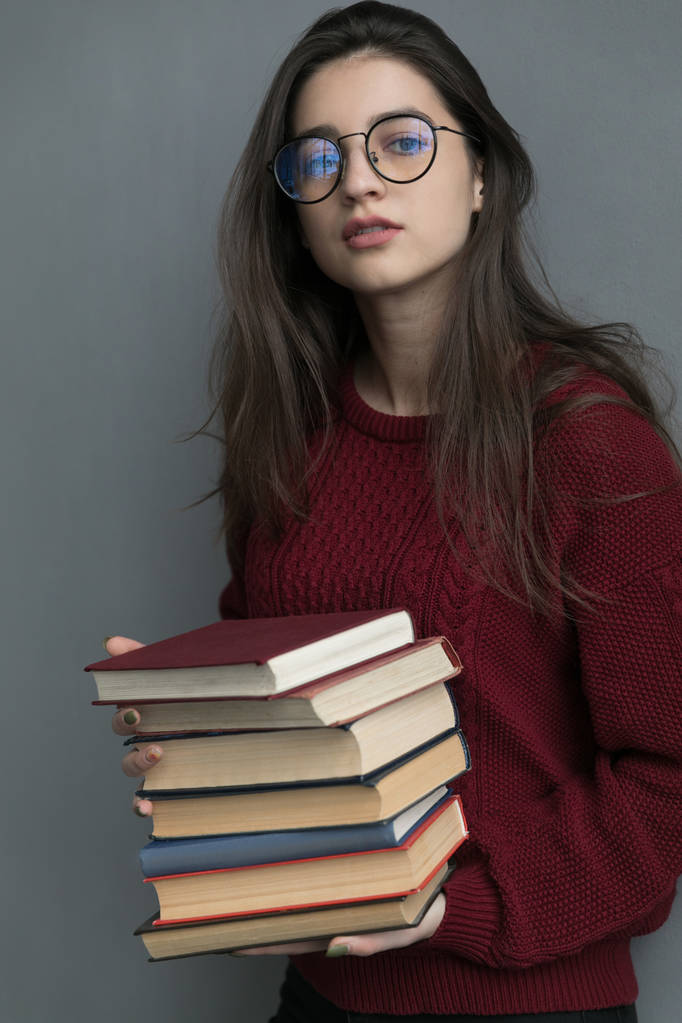 Close-up de uma menina com cabelo fluindo, em um fundo cinza segurando livros em suas mãos, posando. Usando uma camisola e óculos de burgundy. - Foto, Imagem