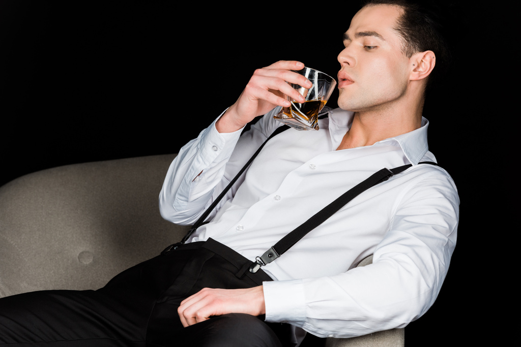 όμορφος άντρας που κοιτάζει το ποτήρι του ουίσκι ενώ κάθεται σε πολυθρόνα απομονώνεται σε μαύρο  - Φωτογραφία, εικόνα