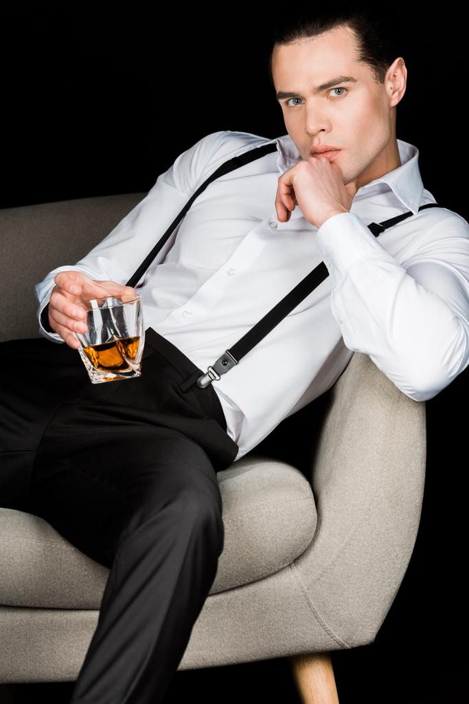 hombre serio sosteniendo vaso de whisky y tocando la cara mientras está sentado en sillón aislado en negro
  - Foto, Imagen