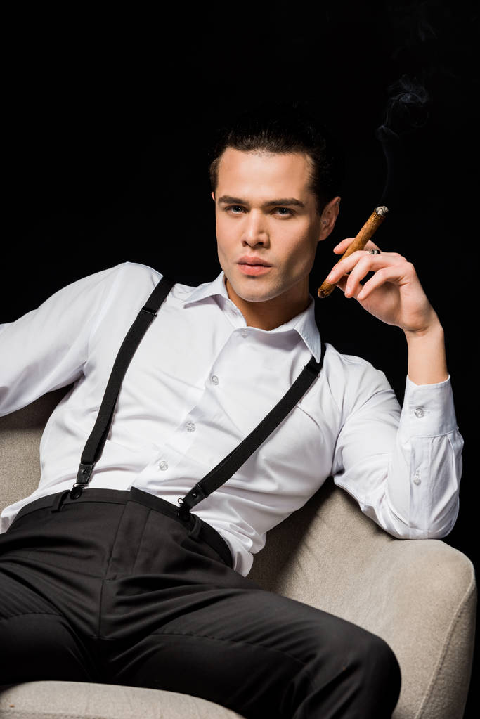 handsome man holding cigar while sitting in armchair isolated on black  - Photo, Image