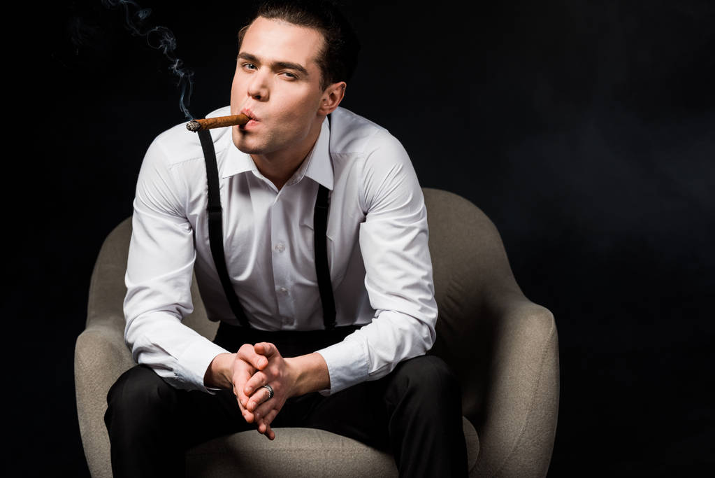 hombre guapo fumar cigarro mientras está sentado en el sillón con las manos apretadas aisladas en negro
  - Foto, imagen