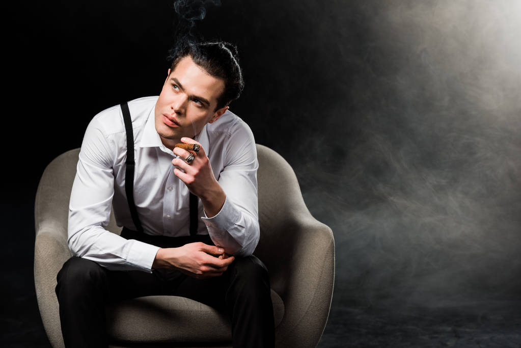 煙で黒い上の肘掛け椅子に座っている間、葉巻を持つ深刻な男 - 写真・画像