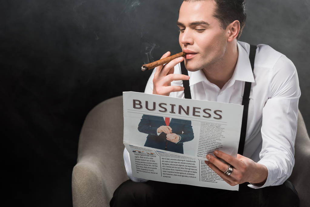 muž sedící v křesle čtení obchodních novin a kuřácký doutník na černém s kouřem  - Fotografie, Obrázek
