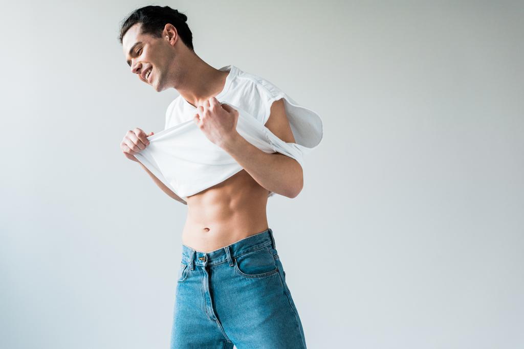 glücklicher muskulöser Mann berührt weißes T-Shirt, während er auf weißem steht  - Foto, Bild