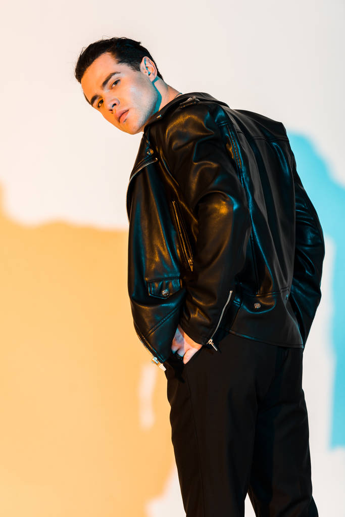 hombre guapo en chaqueta de cuero negro de pie y mirando a la cámara en blanco con iluminación
  - Foto, Imagen