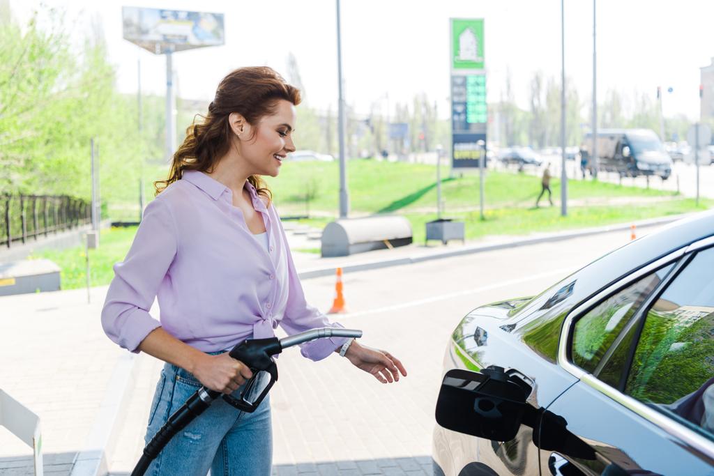 mulher alegre segurando bomba de combustível perto de carro preto no posto de gasolina
  - Foto, Imagem