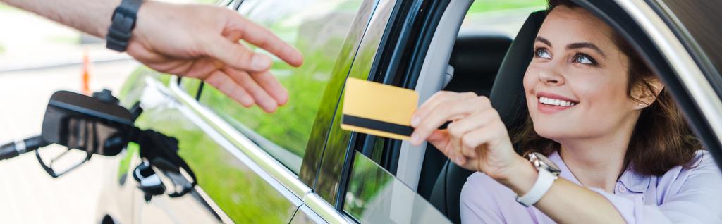 panoramiczny strzał wesoły kobieta siedzi w samochodzie i dając kartę kredytową do pracownika na stacji benzynowej  - Zdjęcie, obraz